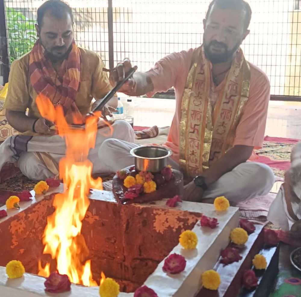 Two Brahmins seated at the fire in Vrindavan, flames rising from the kund, marigolds around it